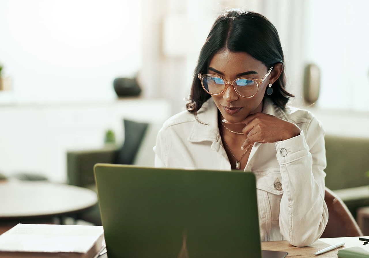 A focused woman in glasses and a white denim jacket is focused on the laptop screen in front of her.