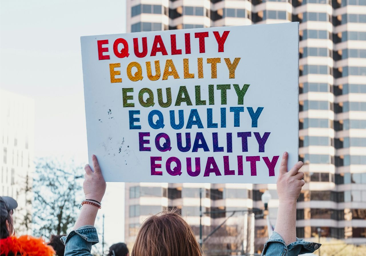 A person holds a sign reading "EQUALITY" in rainbow colors against a backdrop of city buildings.