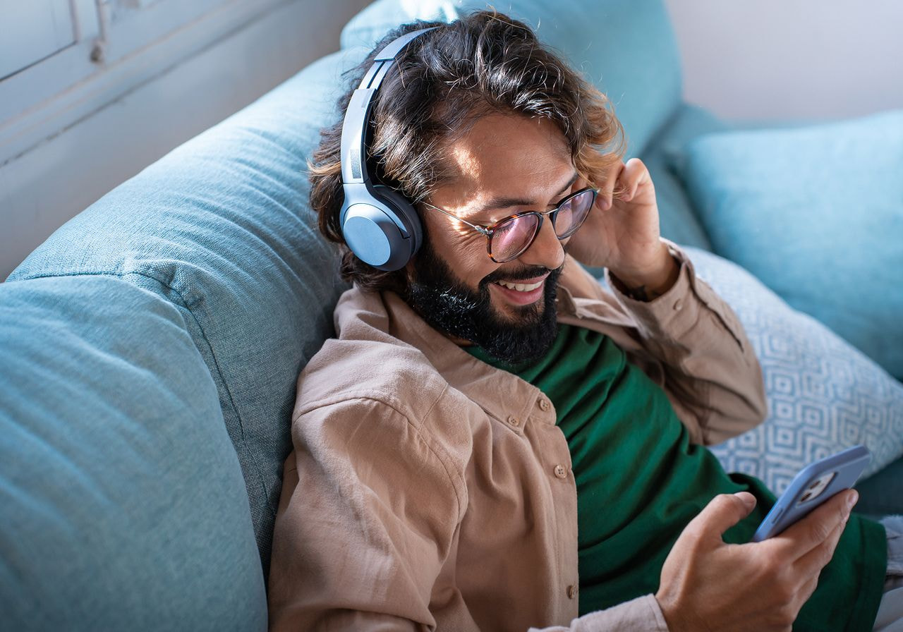 A bearded man wearing headphones sits on a couch, enjoying music in a relaxed setting.