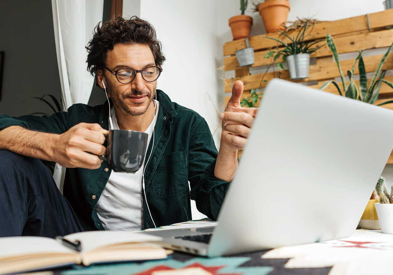 A man wearing glasses, holding a mug, and pointing at a laptop. He is smiling, sitting indoors with earphones.