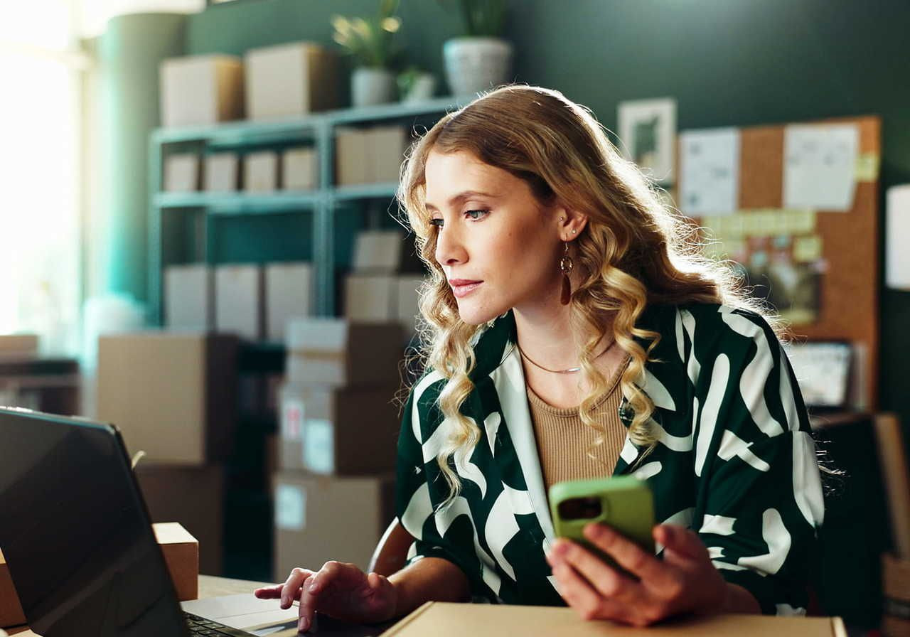 A woman with curly hair is sitting at a desk, looking at a laptop while holding a phone, surrounded by cardboard boxes.