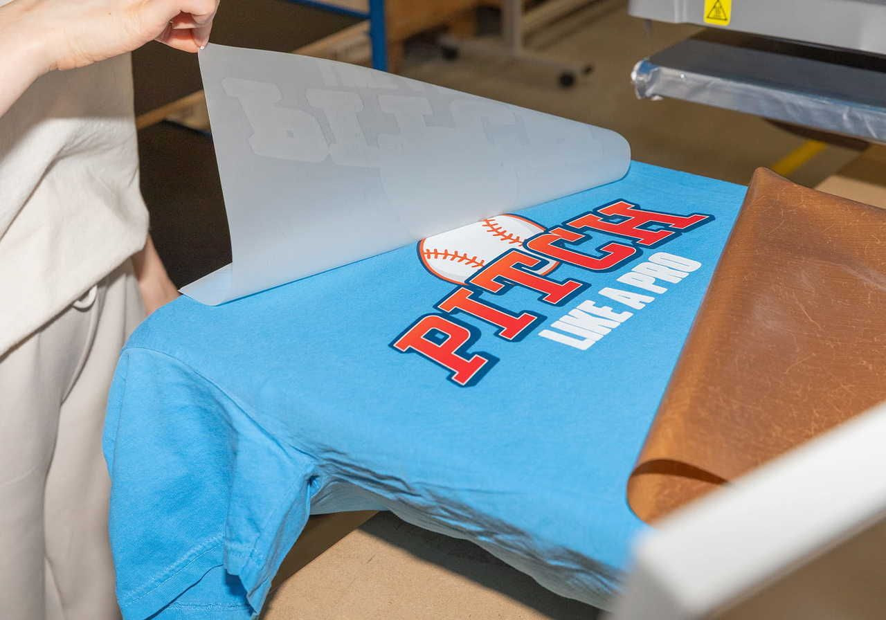 A blue t-shirt with the text "PITCH LIKE A PRO" is freshly printed, with a piece of transfer paper being lifted off.