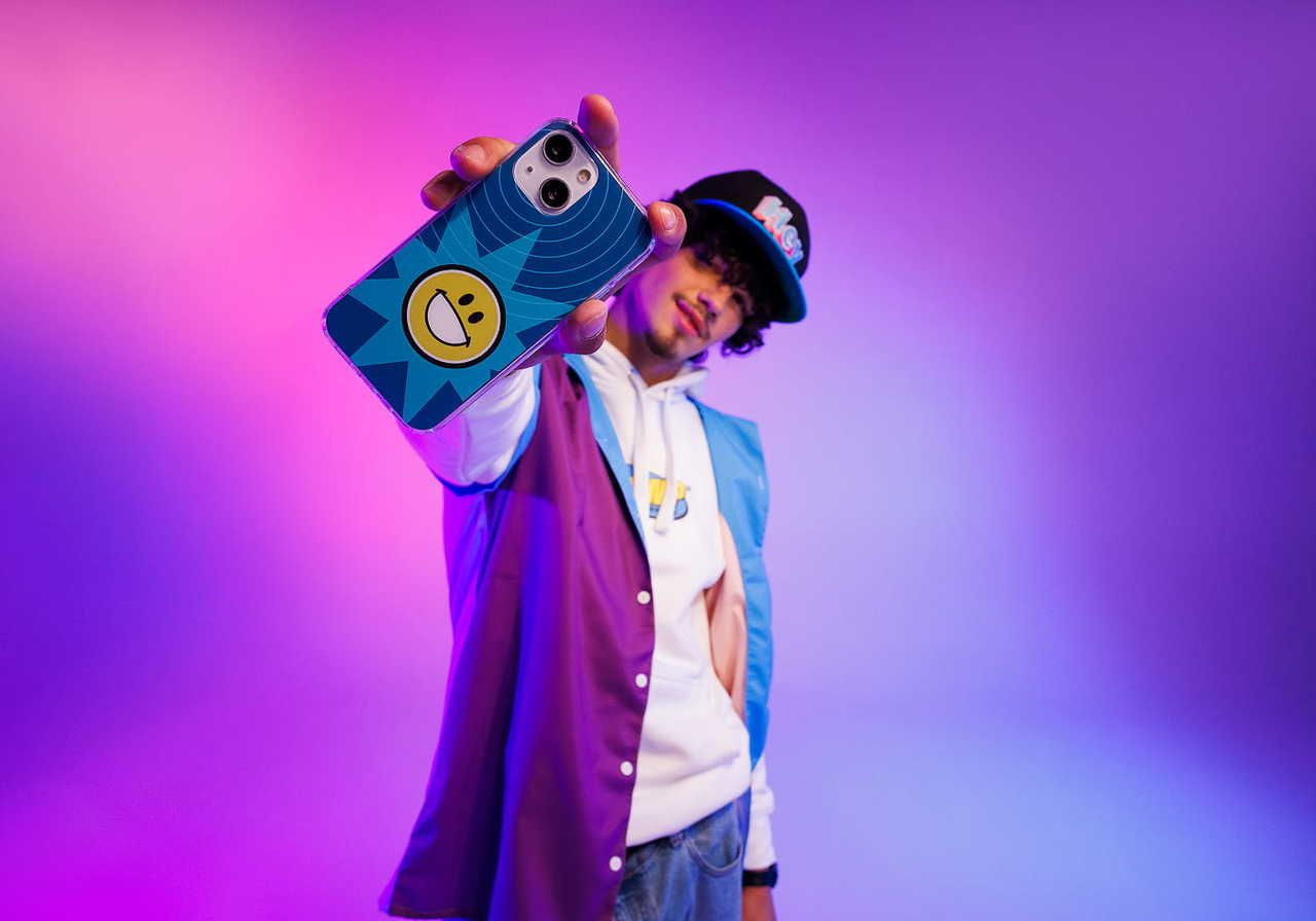 A young man holding a phone with a colorful case, wearing a cap and a stylish outfit, against a vibrant purple and blue background.