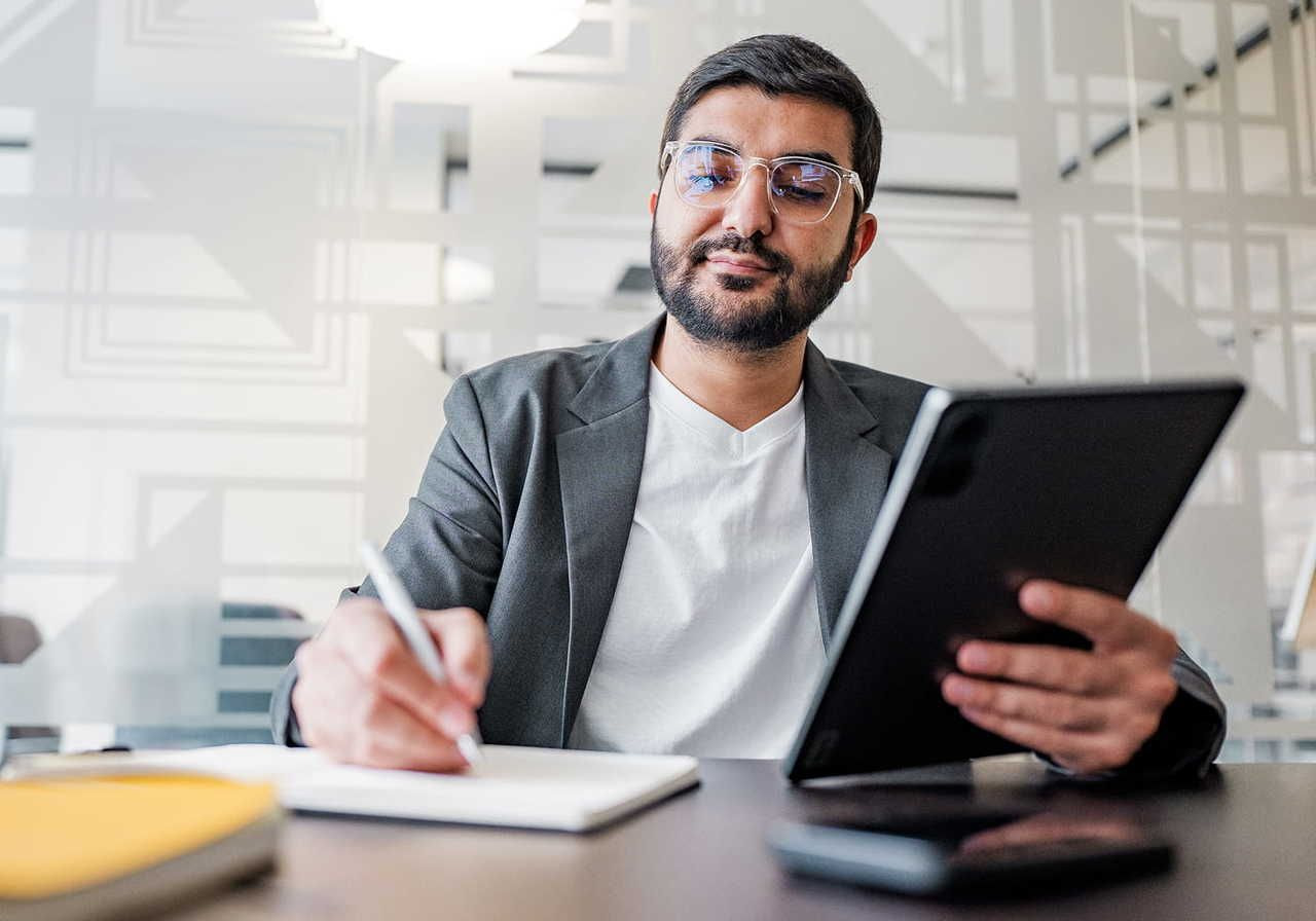 A bearded man in glasses, wearing a blazer in a modern office, holding a tablet and writing in a notebook.
