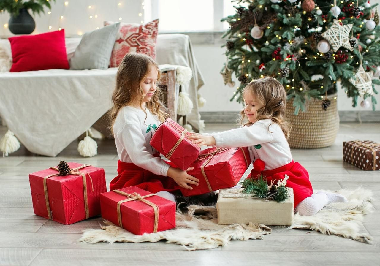 Two little girls in red dresses sit on the floor surrounded by colorful presents, smiling and enjoying the moment.