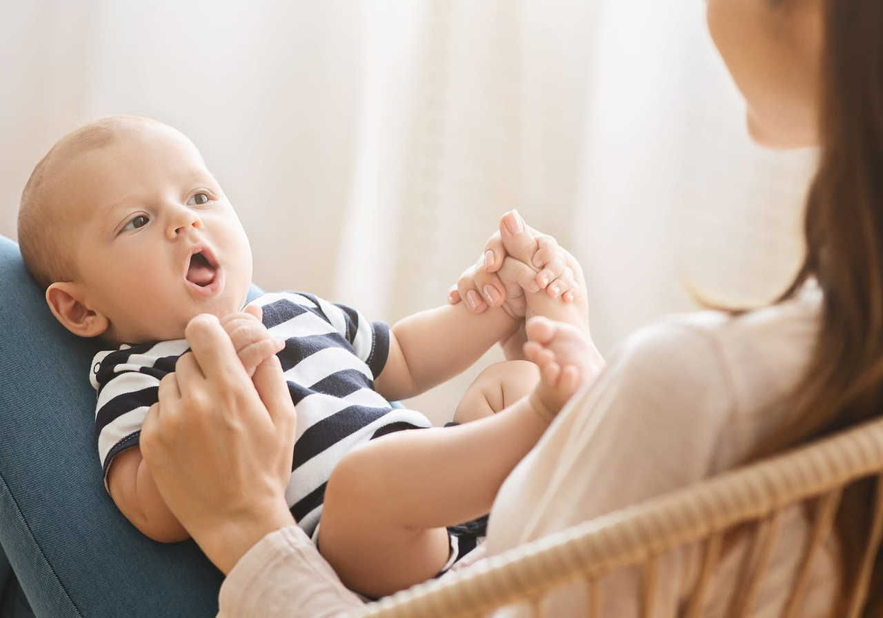 A baby in a black-and-white striped onesie lies in an adult's lap, holding the adult's hand. The baby looks upward with an open mouth.