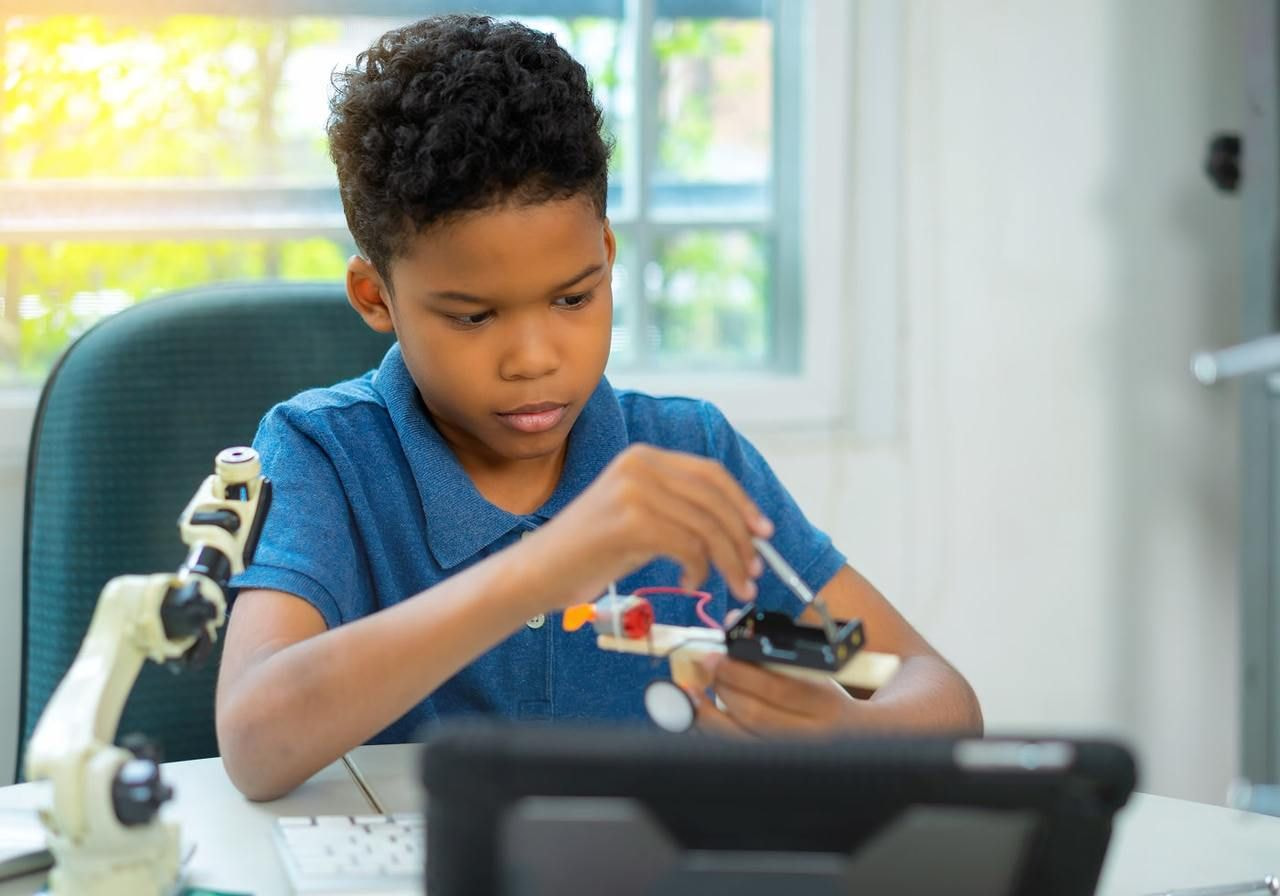 A young boy in a blue shirt focuses intently on building a DIY robot at a desk. A microscope and a laptop are nearby.
