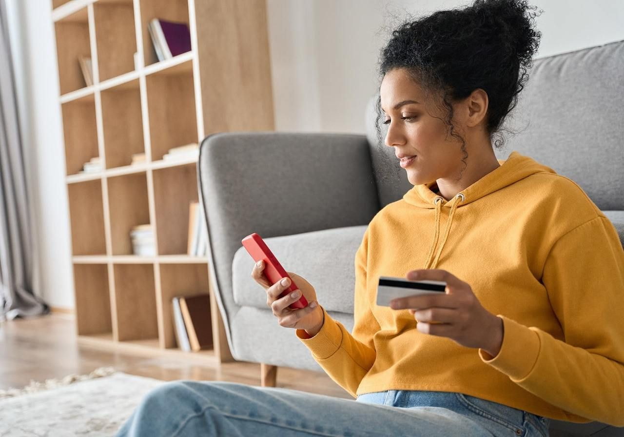 A woman in a yellow hoodie sits on the floor, holding a credit card and smartphone, likely shopping online.