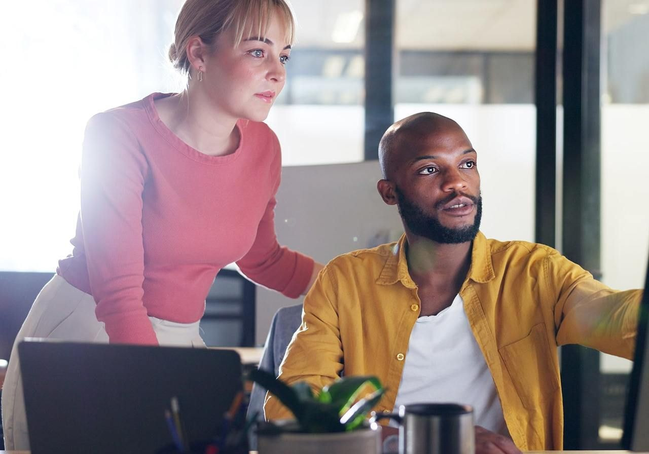 Two colleagues in an office collaborate at a computer, one seated and pointing at the screen, the other standing and observing.