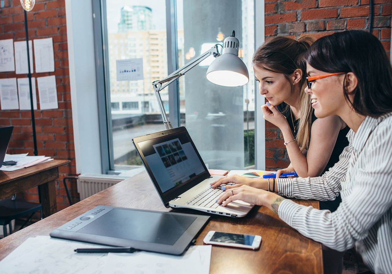 Two women collaborating on a laptop in a modern office setting, focused on their work.