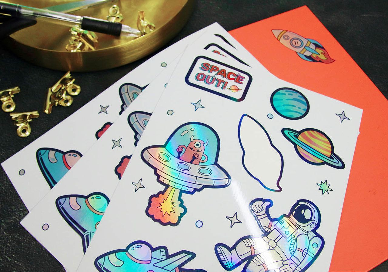 A set of holographic space-themed stickers on a dark surface, featuring rockets, planets, an alien in a UFO, and an astronaut.
