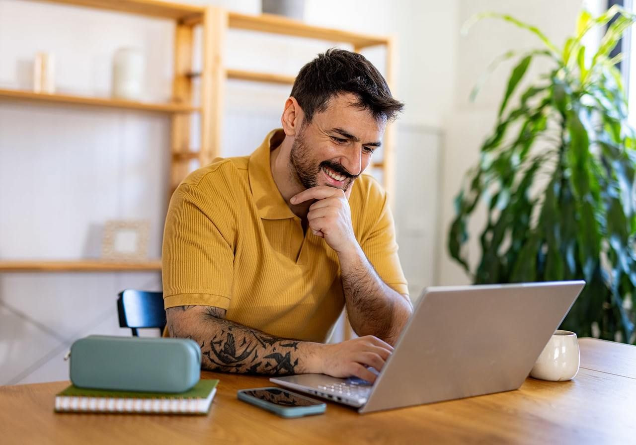 A smiling man in a yellow t-shirt is using a laptop.