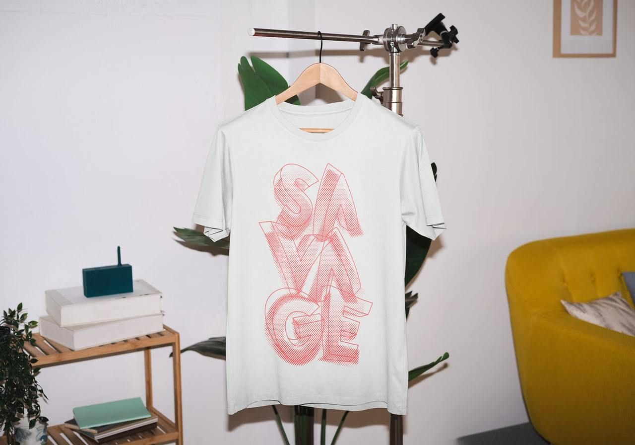White t-shirt on a hanger with bold red letters spelling "SAVAGE." Background includes a yellow chair, a plant, a shelf, and art.