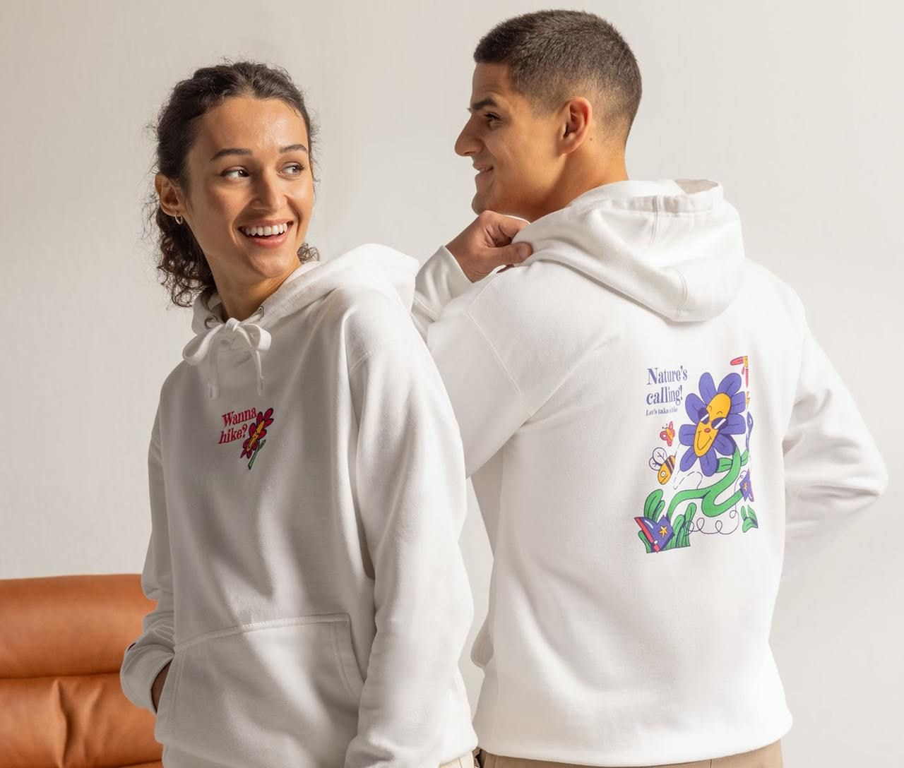 A smiling woman and a man in white hoodies with colorful designs. Her hoodie reads "Wanna hike?". His has a playful flower illustration.