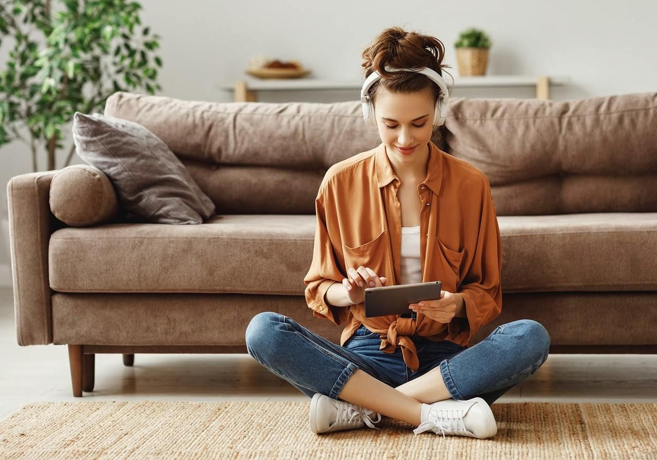 A woman sits cross-legged on a rug, wearing headphones and using a tablet, with a couch and plant in the background.