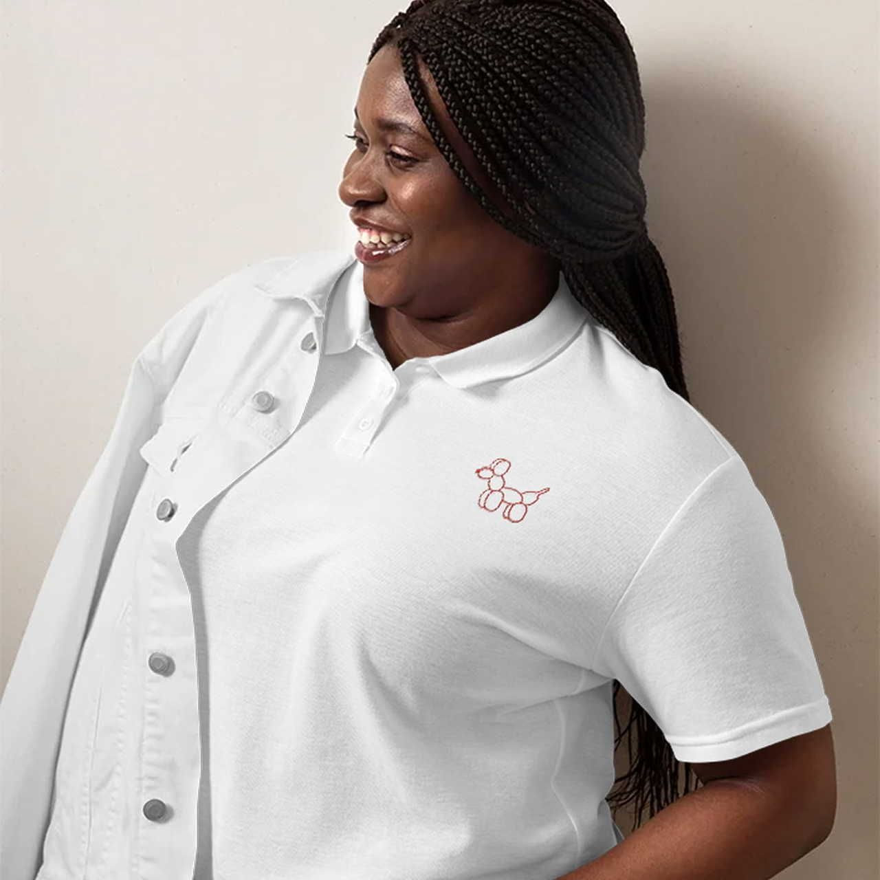 A woman smiling warmly, wearing a white polo with a red balloon dog embroidered logo. She has braided hair and a white jacket.