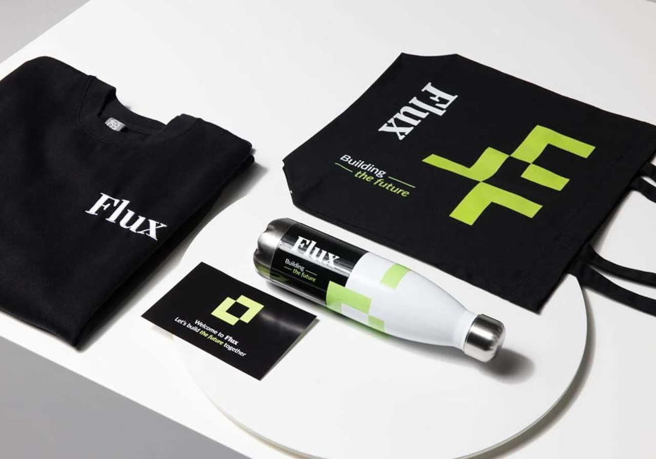 A black t-shirt, a black tote bag with a green logo, a silver bottle, and a card are arranged on a white surface.