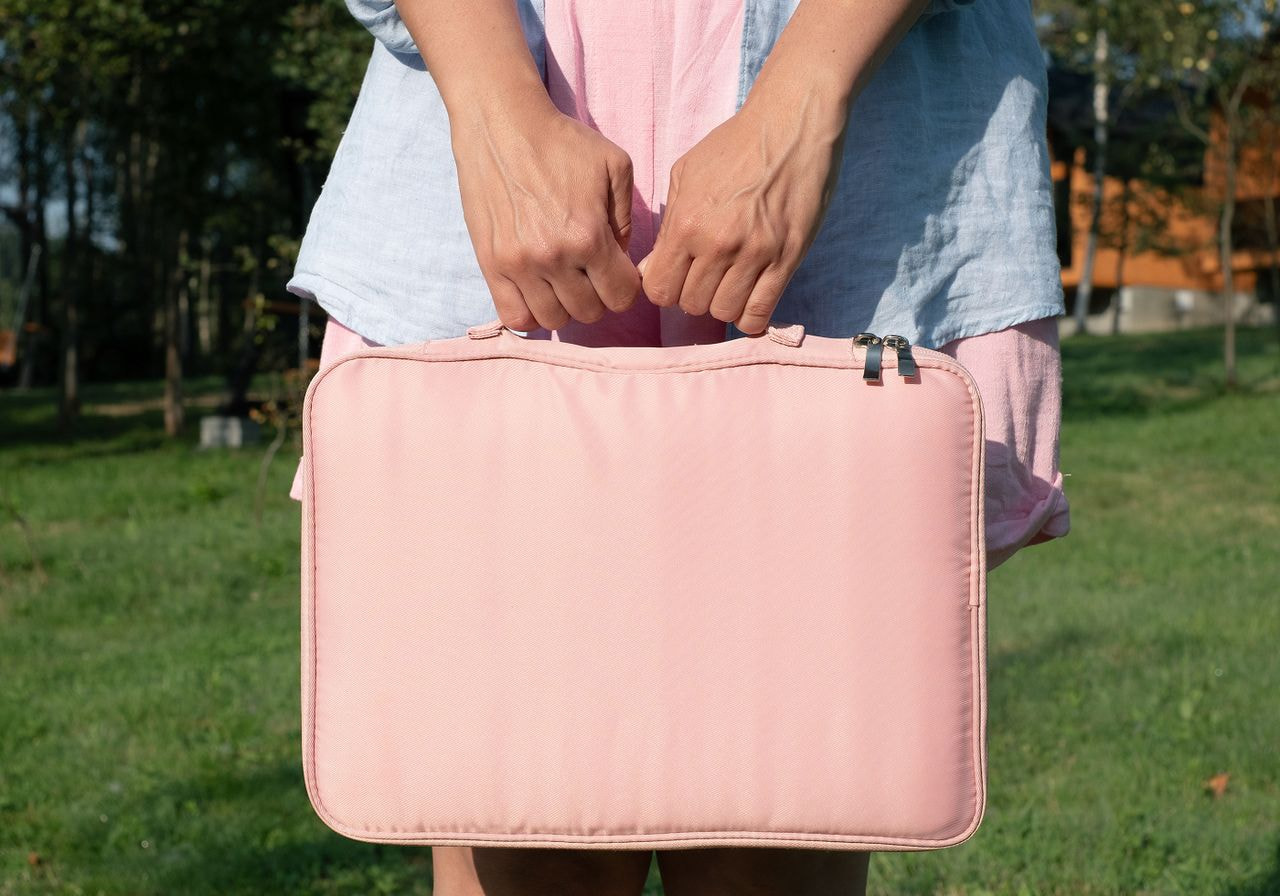 A person holds a blush pink laptop sleeve by its handles while standing on a sunny lawn.