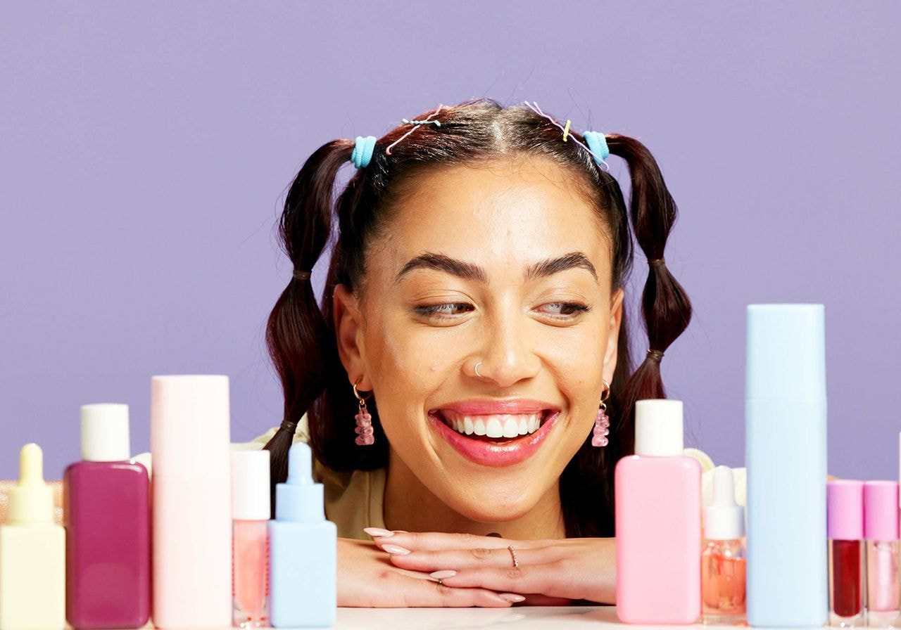 Smiling woman with playful hairstyle and earrings gazes at colorful skincare bottles on a table against a lavender backdrop, conveying a joyful mood.