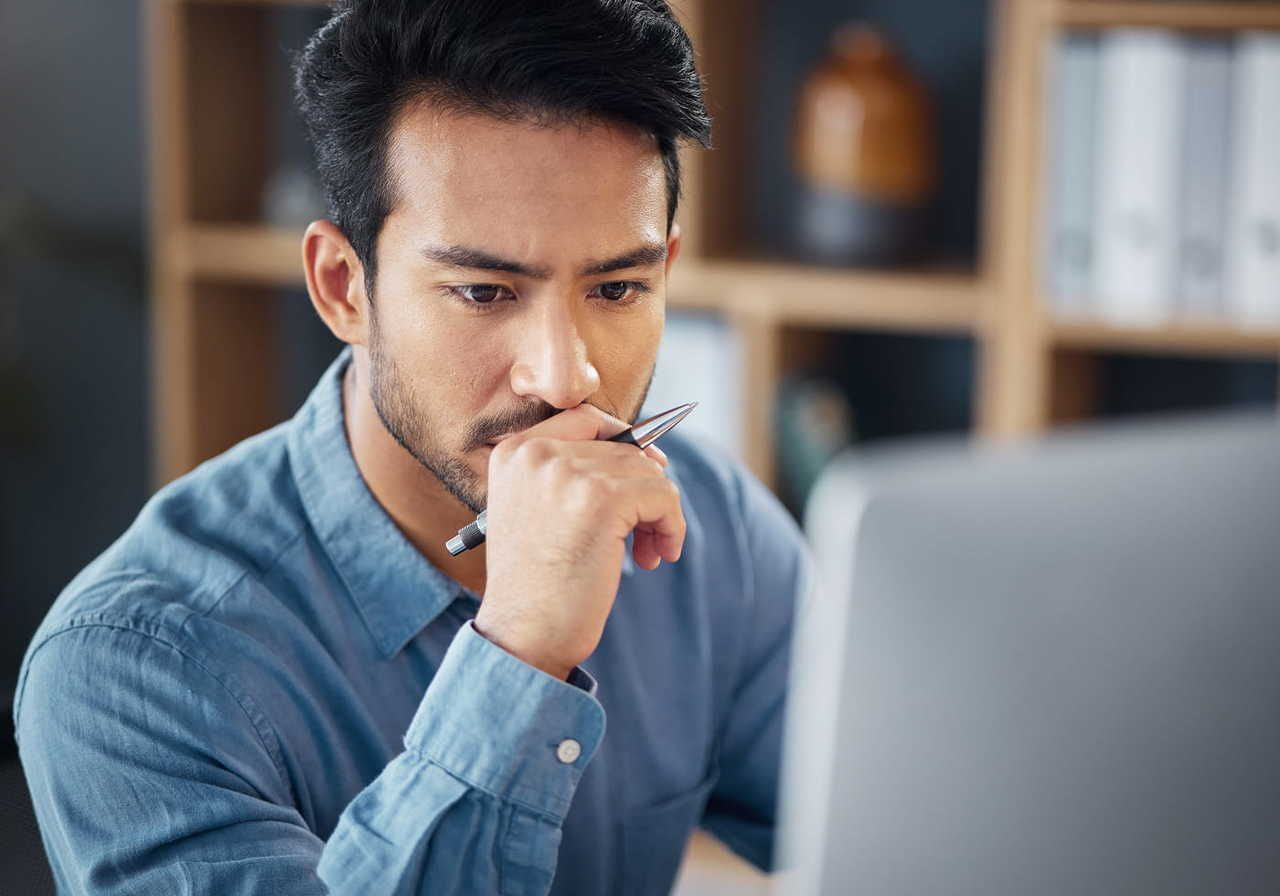 A man in a blue shirt is sitting at a desk, looking thoughtfully at a computer screen while holding a pen.