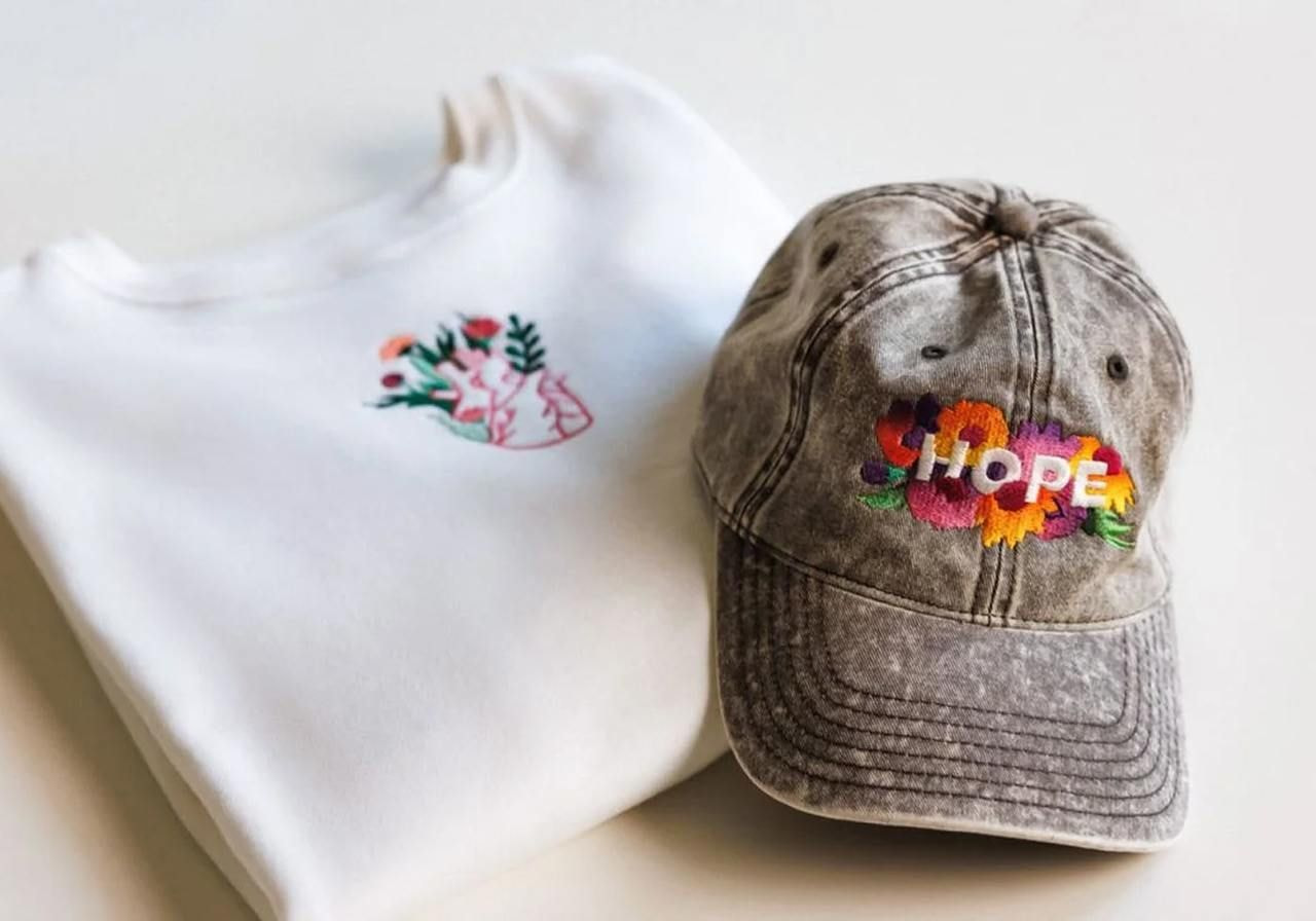A white sweatshirt with floral embroidery next to a faded gray cap featuring colorful flowers and the word "HOPE."