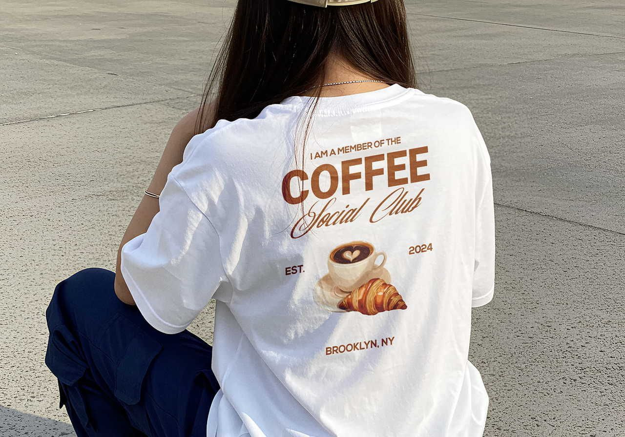 A woman wearing a white "Coffee Social Club" t-shirt with a coffee cup and croissant design sits on concrete.