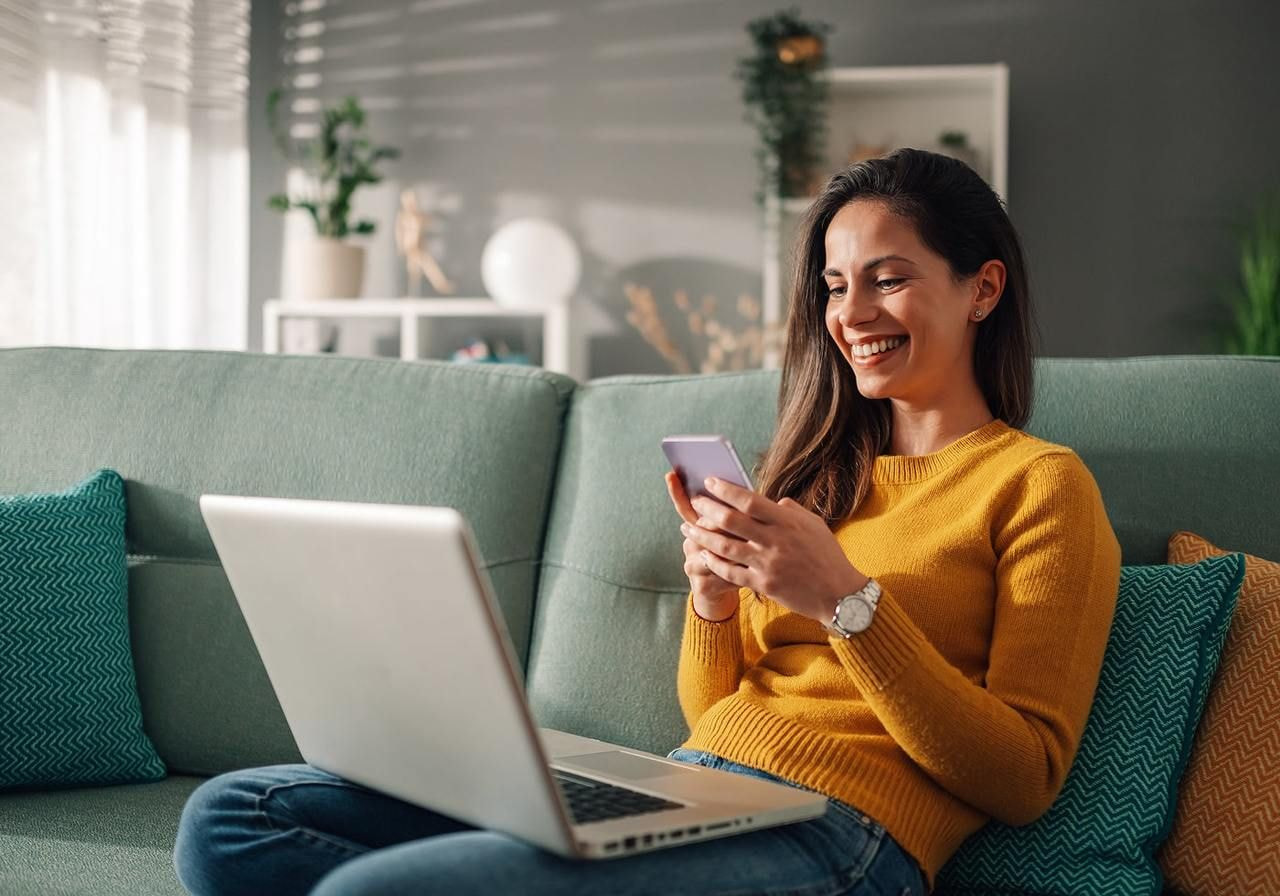 A woman in a yellow sweater sits on a green sofa, smiling at her phone. A laptop rests on her lap.