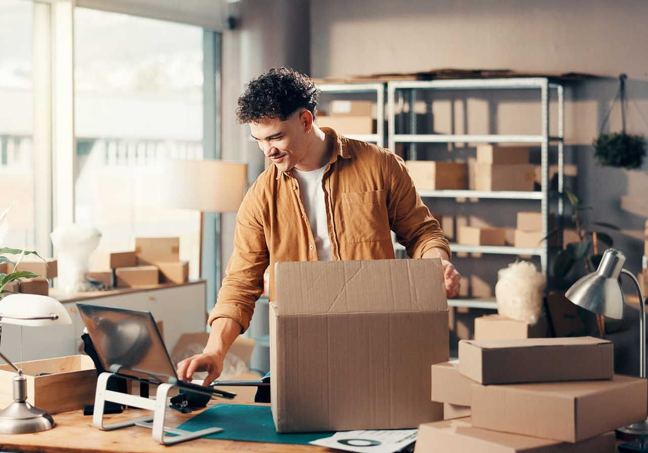 A smiling man is typing on a laptop with one hand while closing a cardboard box with the other. Shelves and multiple boxes in the background.