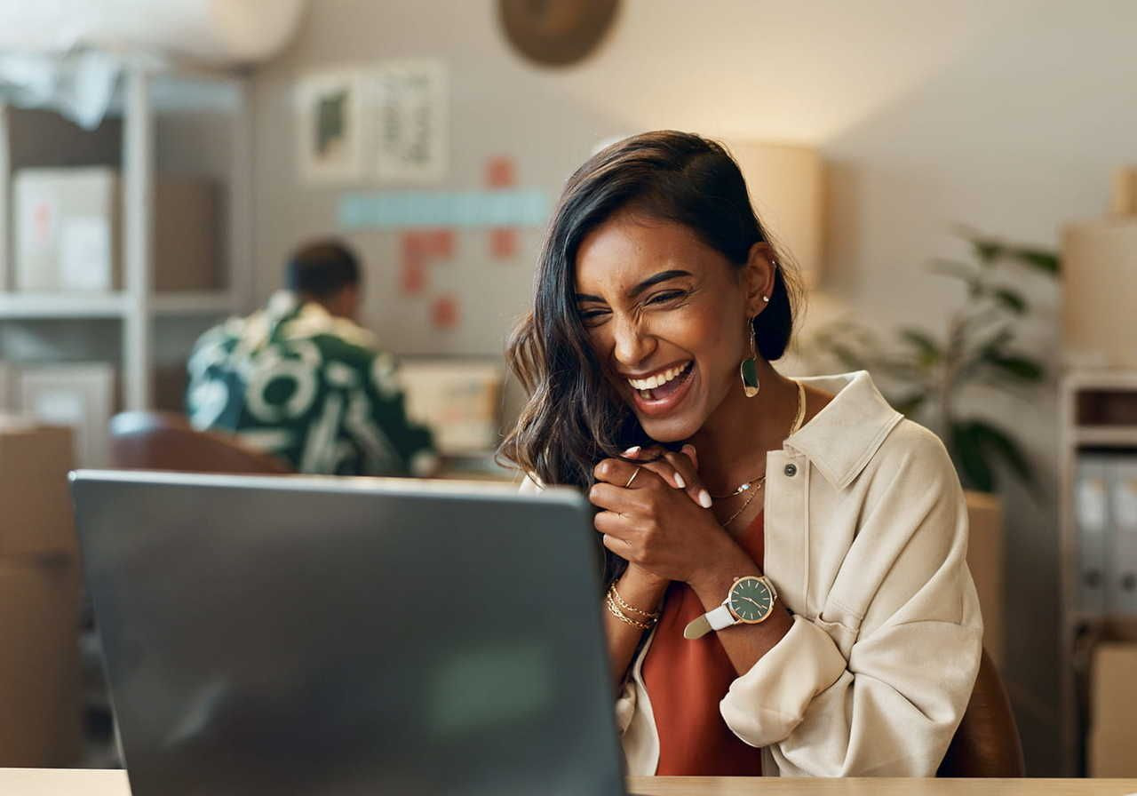A woman laughing joyfully while sitting in front of a laptop in a cozy office environment.
