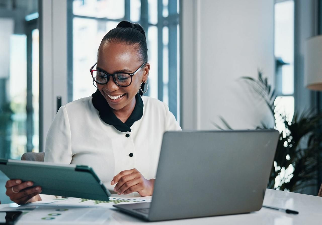 A smiling woman is sitting at a desk in front of a laptop and using a tablet.