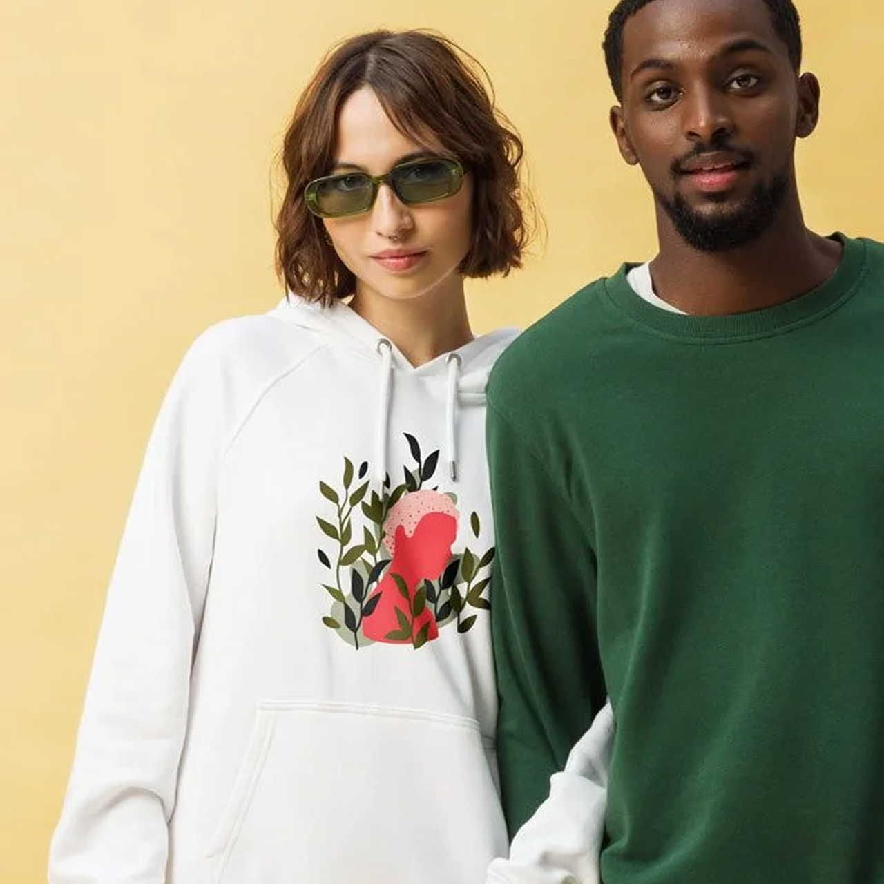 Two people stand closely against a yellow background. The woman wears sunglasses and a white hoodie with a floral design.