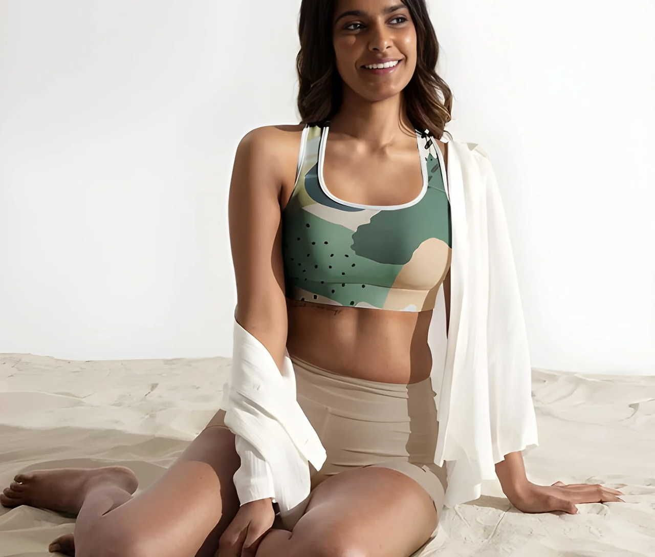 A woman wearing a green patterned sports bra and beige high-waisted shorts, sitting on a sandy surface.