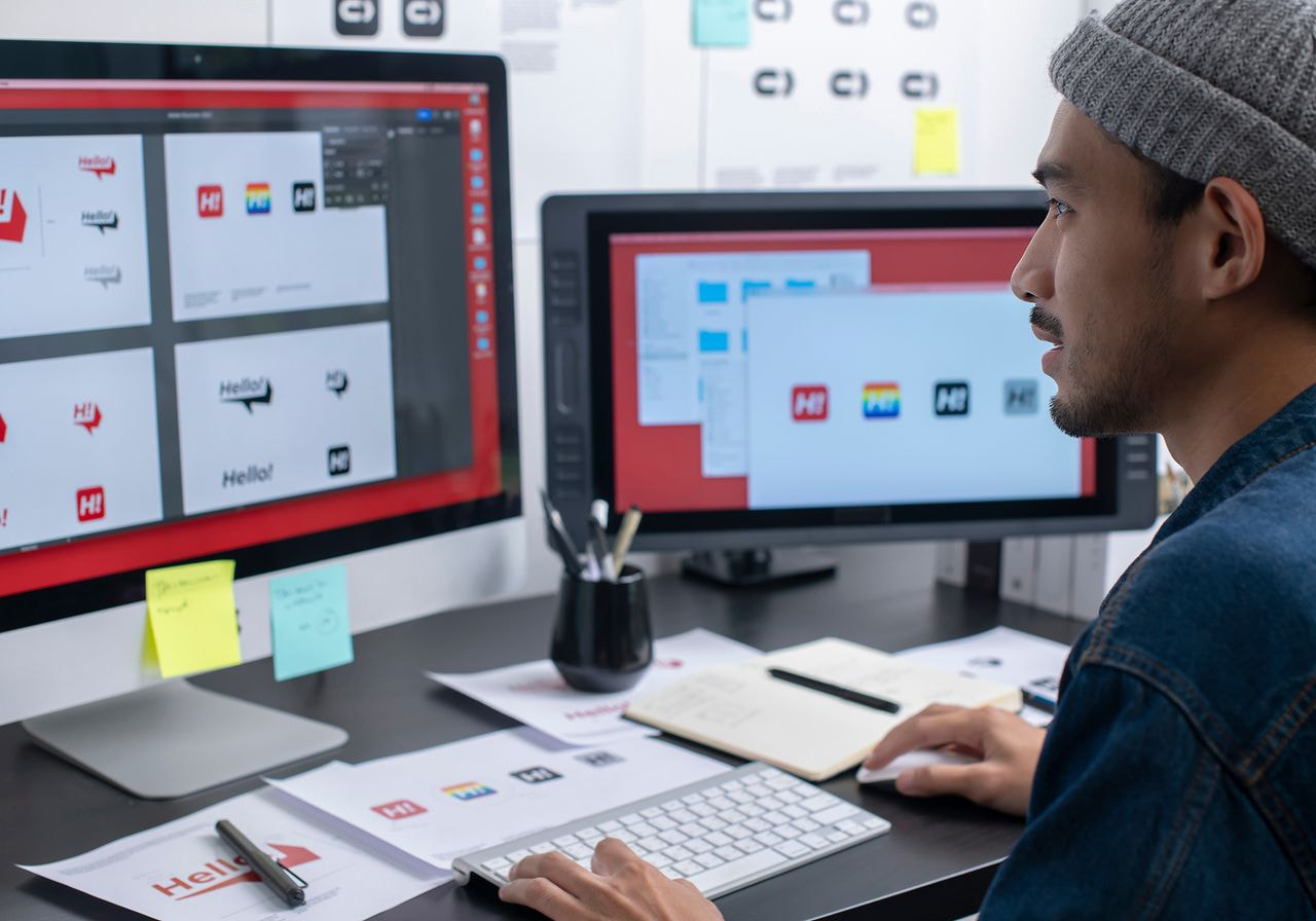 A designer in a beanie reviews logo variations on two computer monitors at a desk covered with printed brand mockups and sticky notes.