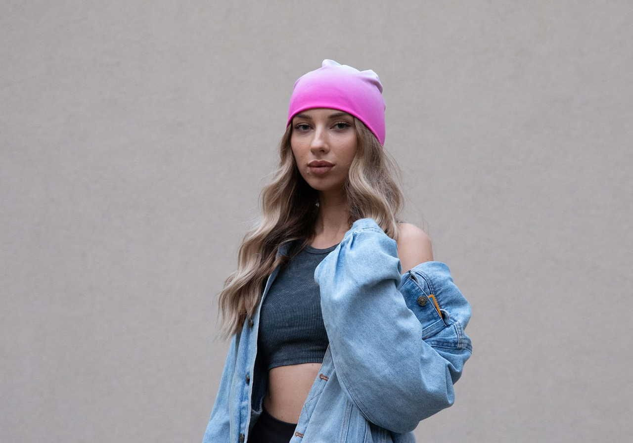 A person with long, wavy hair wearing a pink beanie, a cropped top, and an oversized denim jacket, posing in front of a plain background.