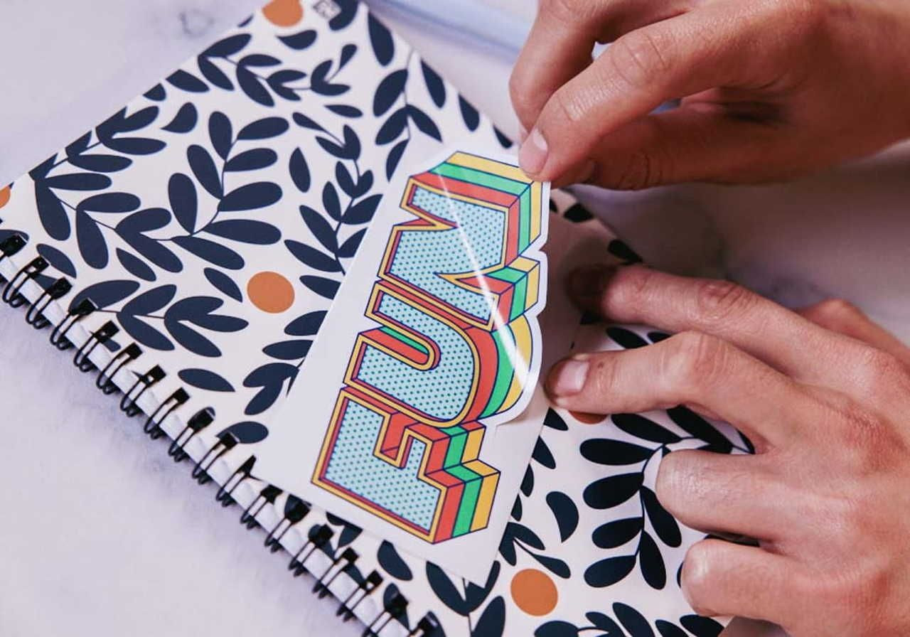 A person placing a colorful "FUN" sticker on a patterned spiral notebook.