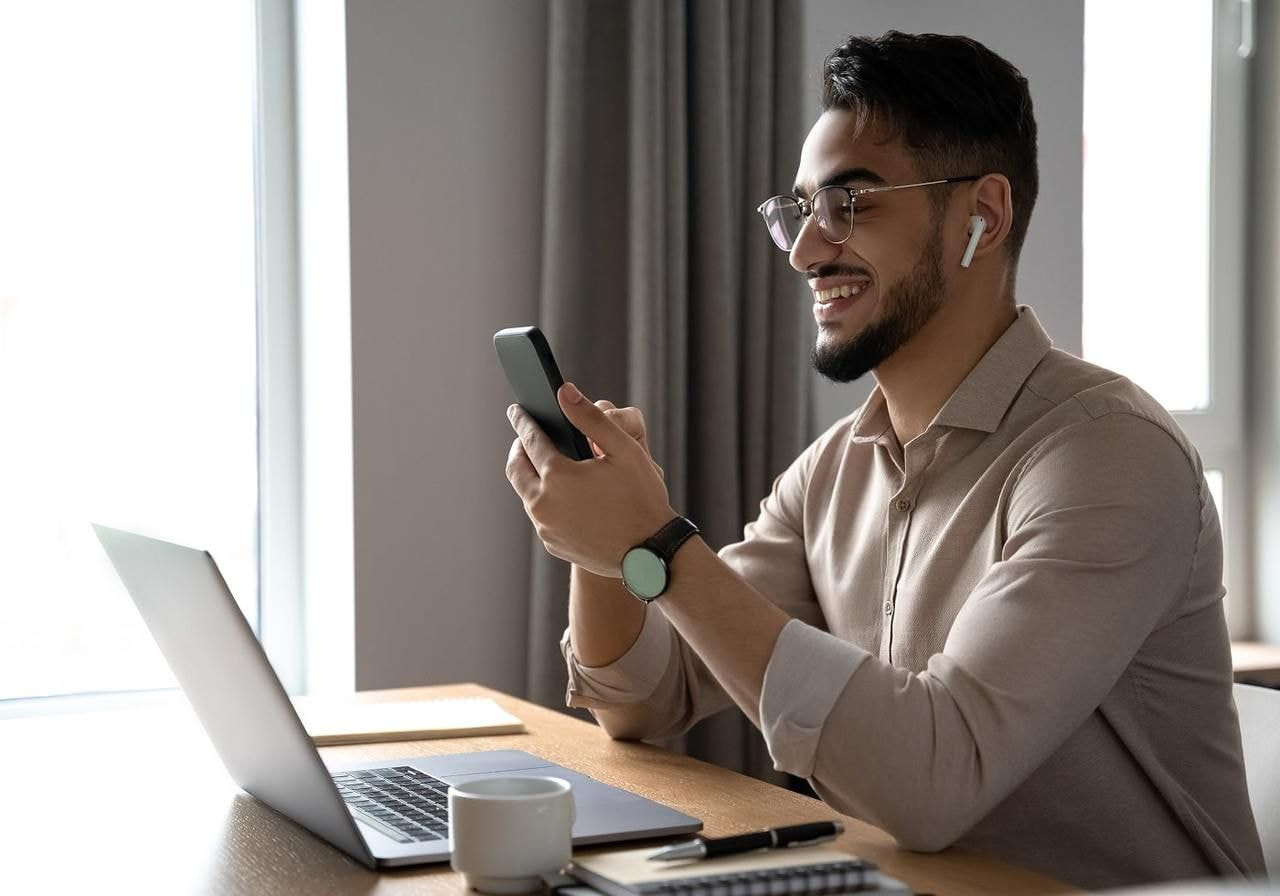 A smiling man with glasses and earphones looks at his phone while sitting at a desk with a laptop, notebook, and mug.