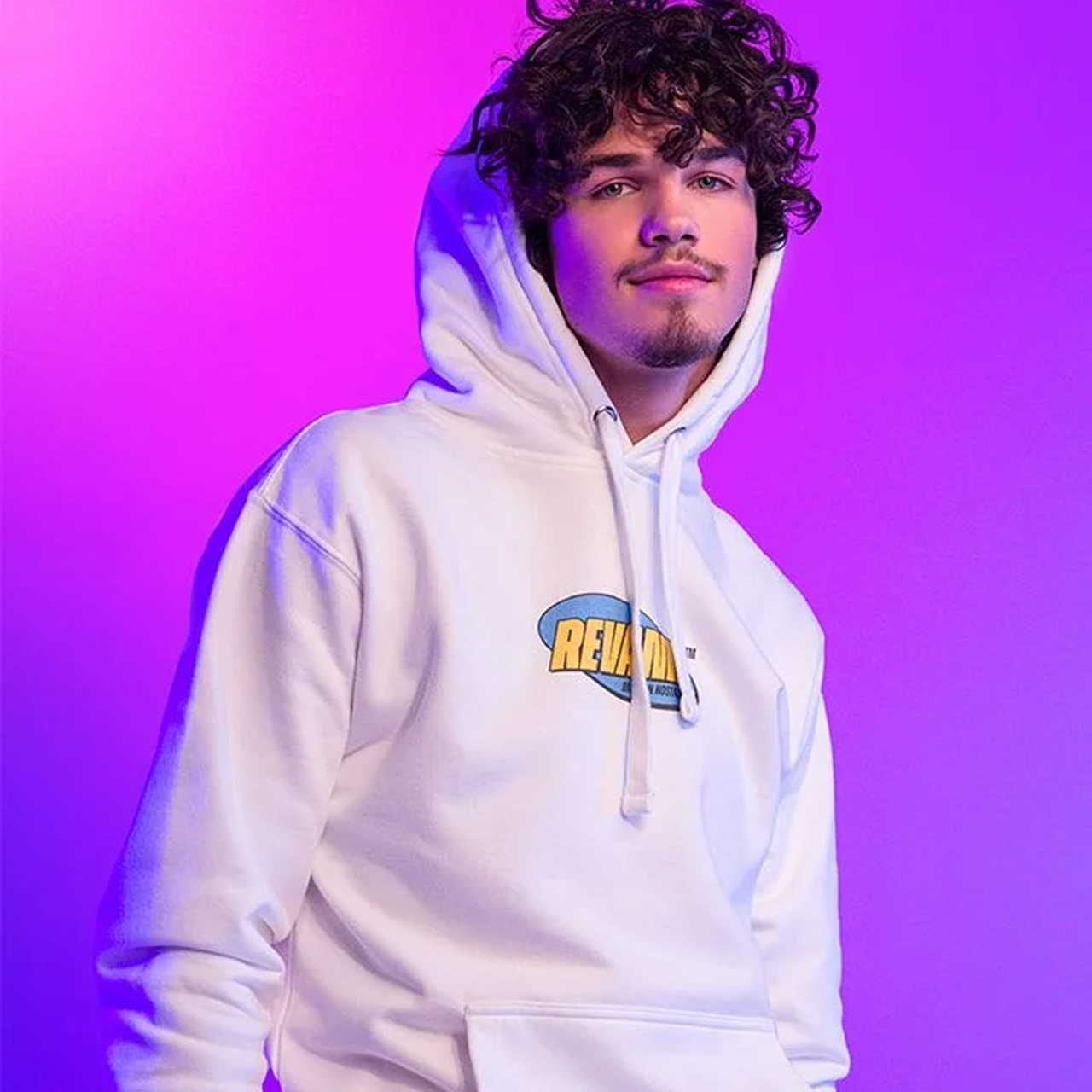 A young man with curly hair wearing a white hoodie featuring a graphic logo stands confidently. Purple and pink lighting background.