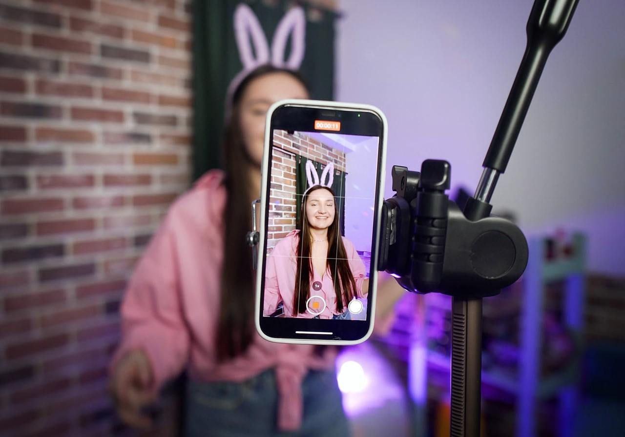 A woman in a pink outfit and bunny ears is recording a video with a smartphone on a tripod.