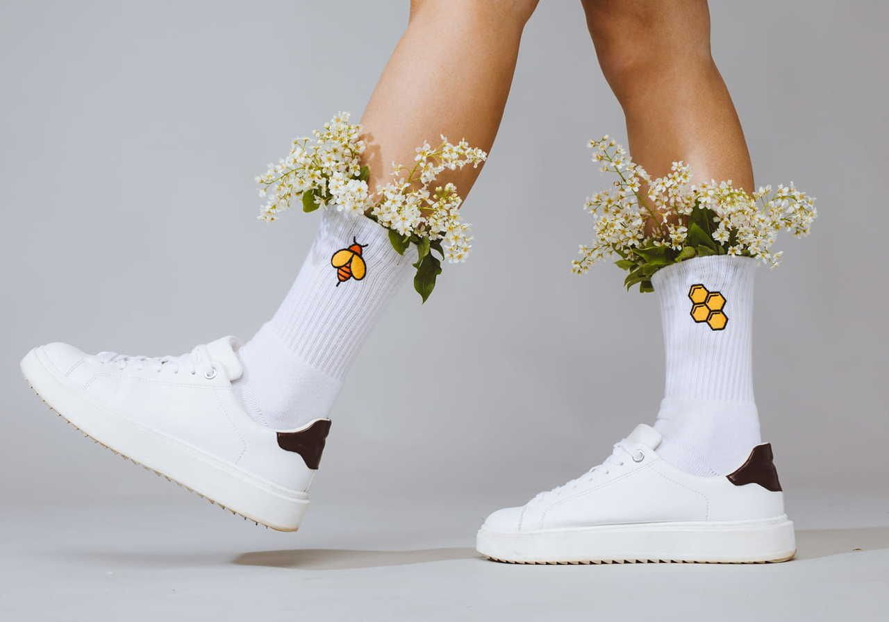 A person wearing white socks with an embroidered bee on one sock, and honeycombs on the second, with small flowers tucked inside.