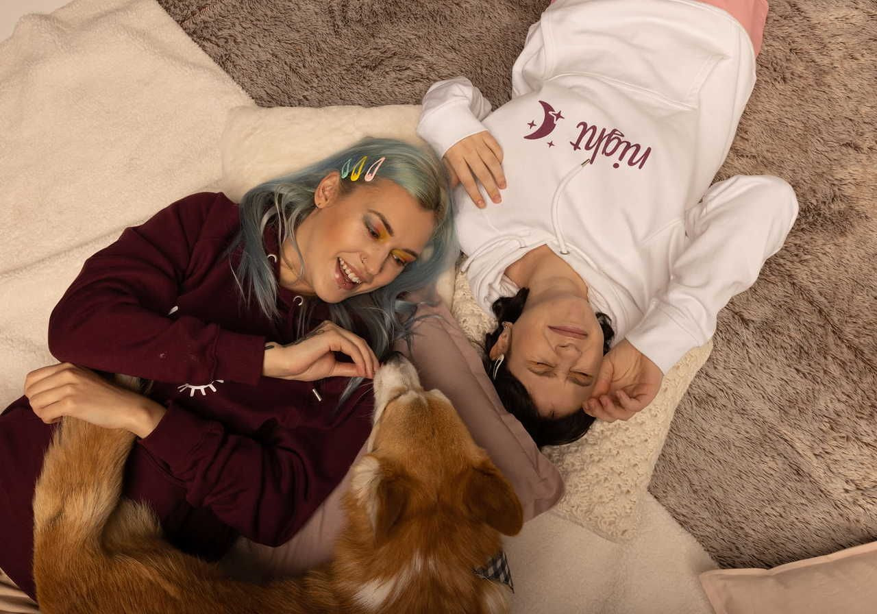 Two young women, one wearing a purple hoodie, second wearing a white hoodie, on a soft, textured surface, smiling and interacting with a dog.