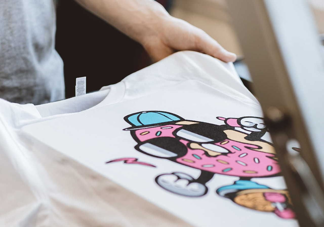 A person taking out a white t-shirt featuring a colorful cartoon character design of a donut with sunglasses and a cap from a DTF printer.