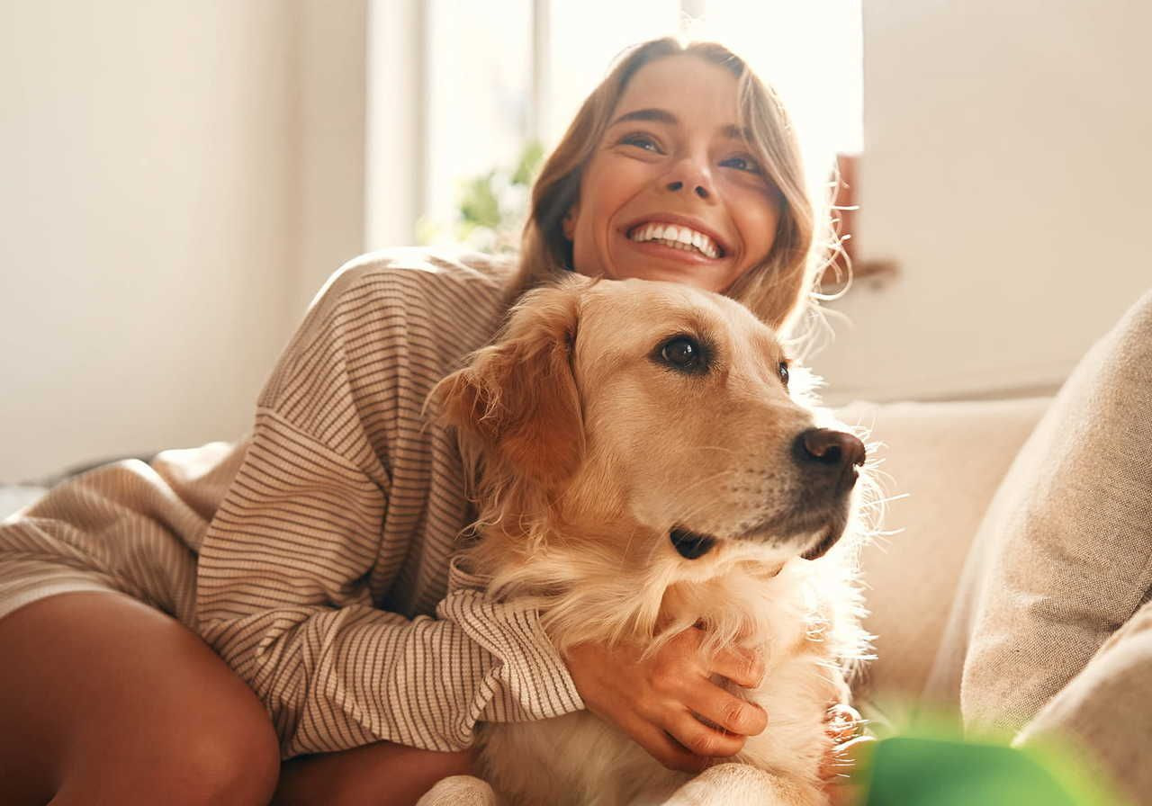A joyful woman with long hair smiles widely while hugging a calm, golden retriever on a cozy couch.