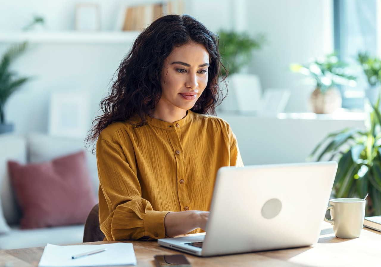 A woman with long, curly hair sitting at a desk, working on a laptop in a bright, modern workspace with plants and soft furnishings.