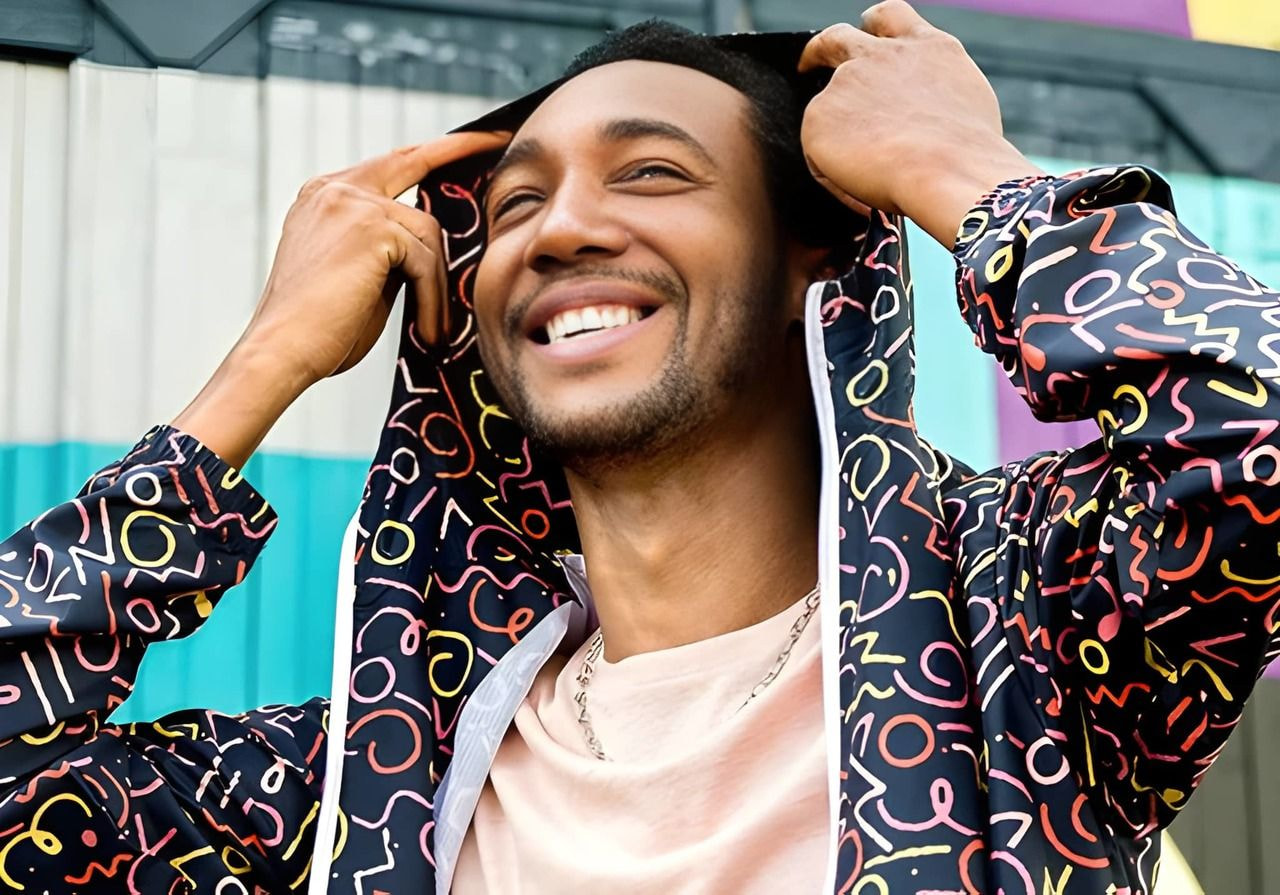 Man smiling joyfully, wearing a windbreaker with colorful abstract patterns. Background features vibrant, striped walls.
