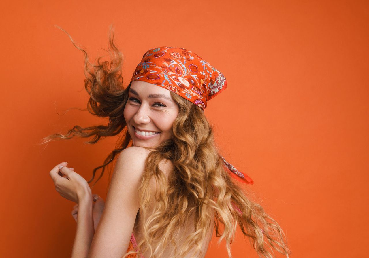 A woman with long wavy hair wears a vibrant orange bandana, smiling joyfully against a matching orange background, exuding a cheerful and lively mood.