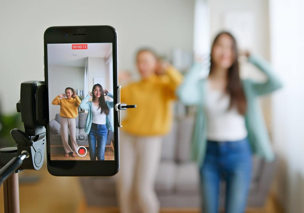 Two women dance happily in a living room, being recorded on a smartphone on a tripod.