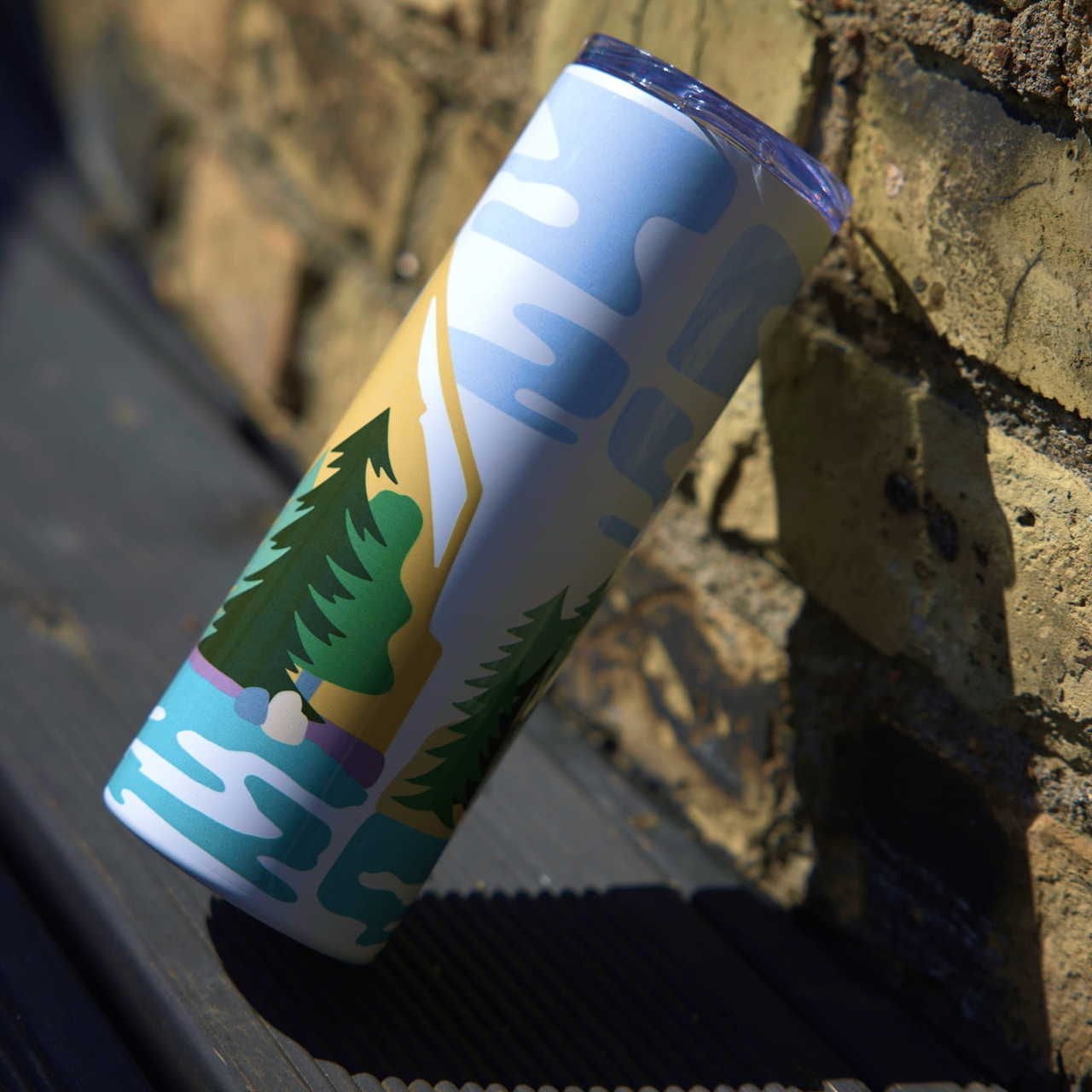 A tall, cylindrical tumbler with colorful outdoors-themed art, including trees and mountains, leans against a textured brick wall.
