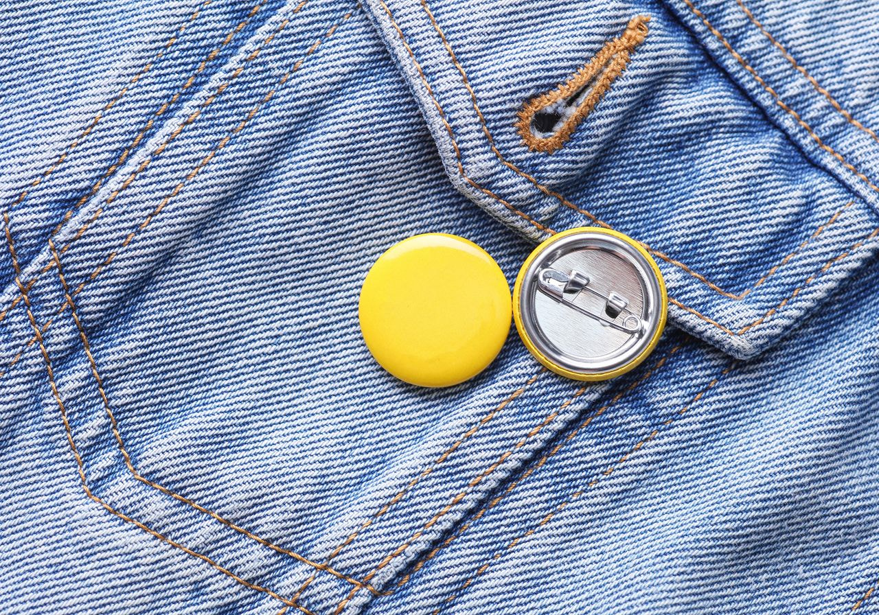 Two blank yellow pin buttons, one face up and one showing its metal clasp, rest on a denim jacket pocket.
