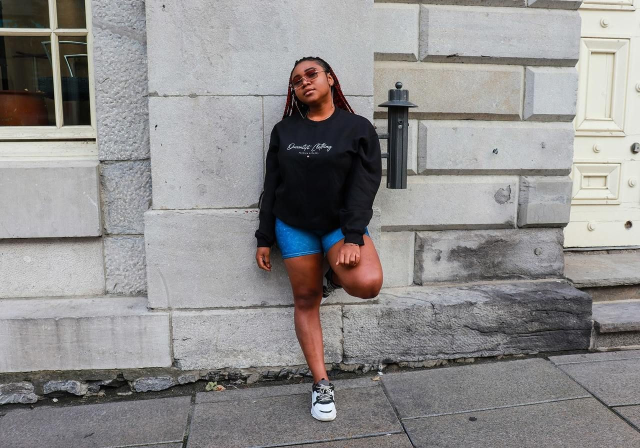 A woman in a black sweatshirt with a chest text print standing in front of a grey wall.