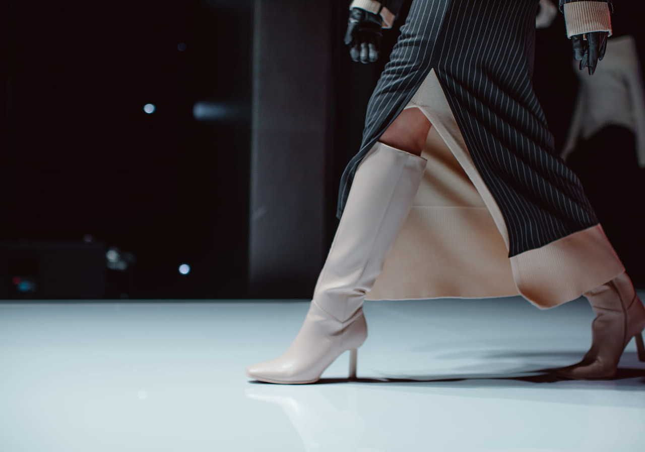 A model in a black pinstripe skirt with a high slit walks confidently in beige knee-high boots on a reflective runway.